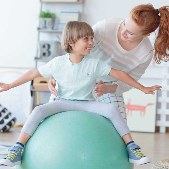 Physical therapist assisting little boy sitting on gym ball during rehabilitation in children hospital